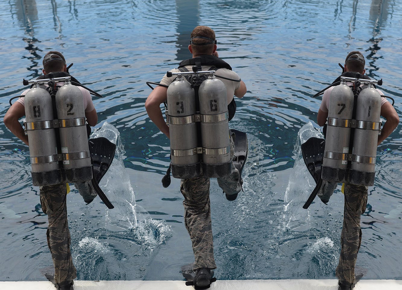 Back view of three divers all about to step into water below them. Each diver has a twin air cylinder; each cylinder has a number stencilled on it.