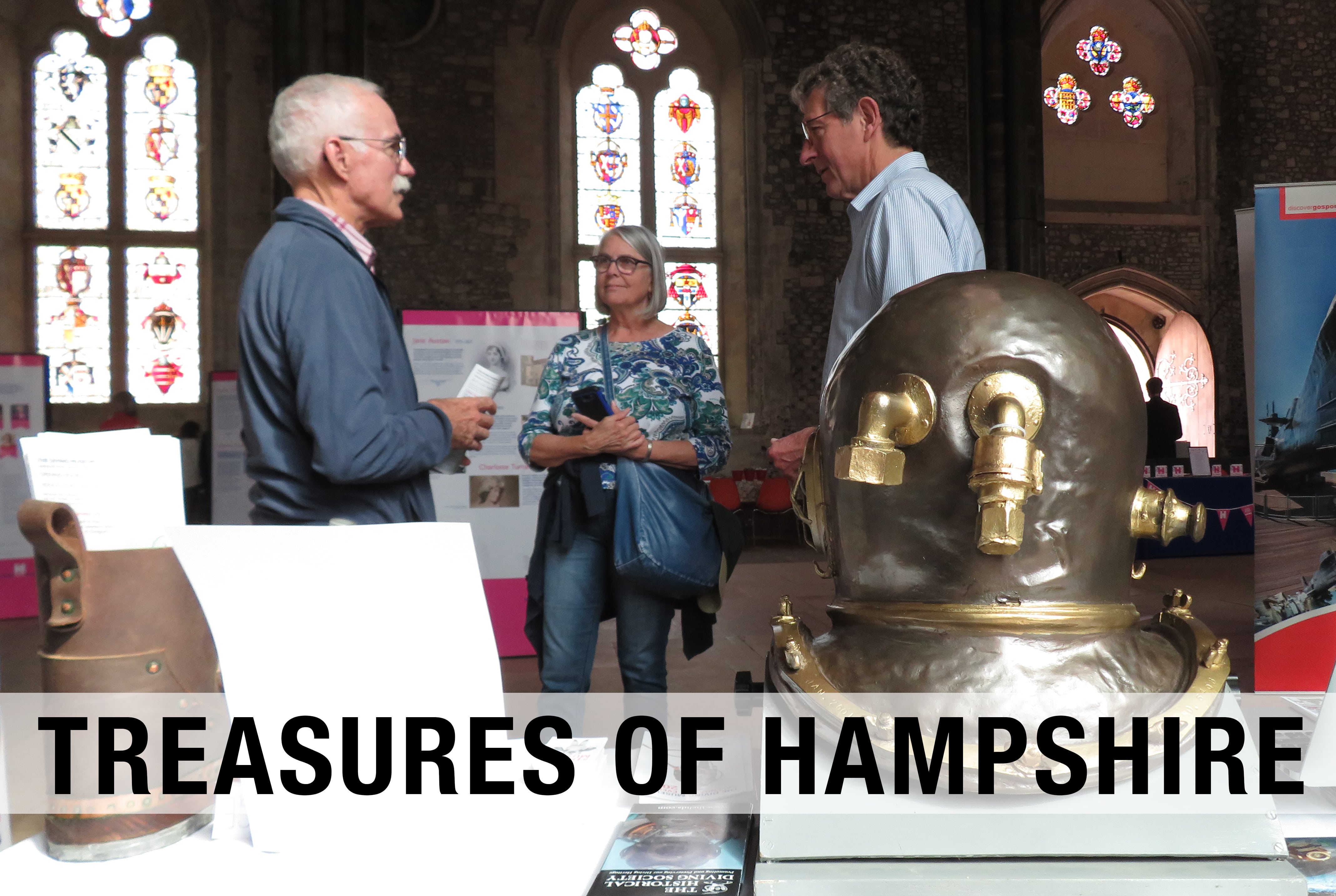 Photograph of three people talking together. They are standing behind a table with a diving helmet on it. The stained glass windows of the Great Hall can be seen in the background.