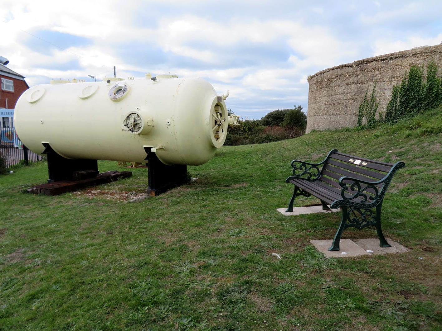 The grassed outdoor area of the museum with a yellow diving chamber on the left and concrete gun emplacement on the right. Between the two is a garden bench.