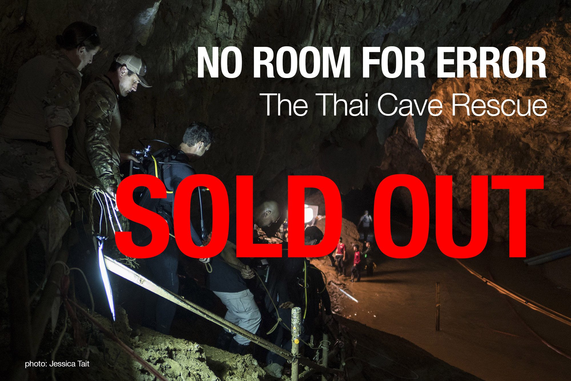 View from high level looking into a sandy-bottomed underground cavern. 5 people including a diver are descending steps to the bottom of the cave where a group of rescuers are gathered around a powerful light 
