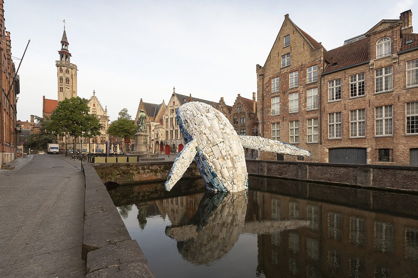 Whale made of plastic waster erupting from a canal in Bruges. We see the white underbelly arching backwards over a pedestrian bridge over the canal with buildings of old Bruges in the background.