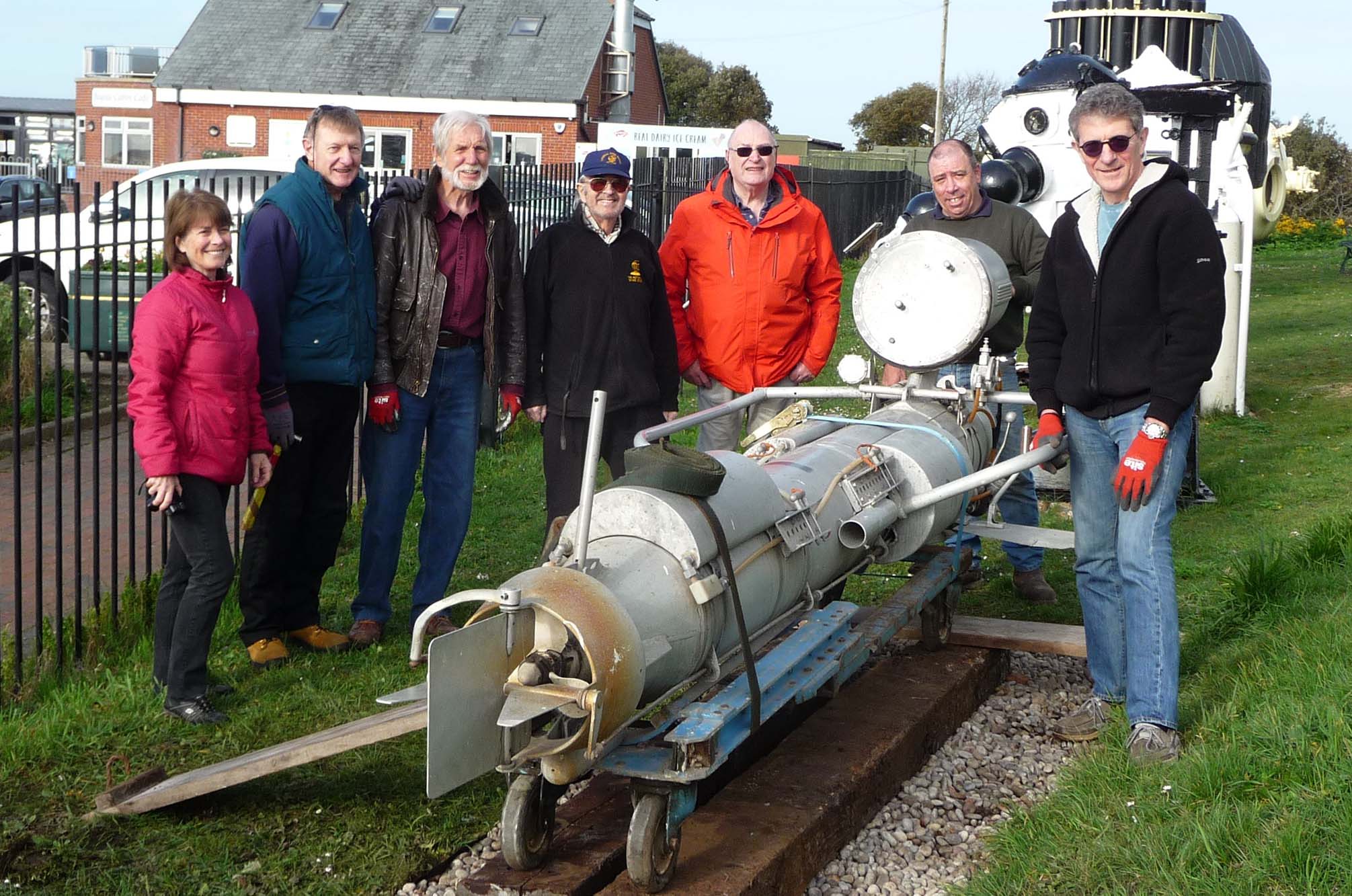 Group of seven museum volunteers standing behind the torpedo-shaped swimmer delivery vehicle in the outside exhibit area of the museum.