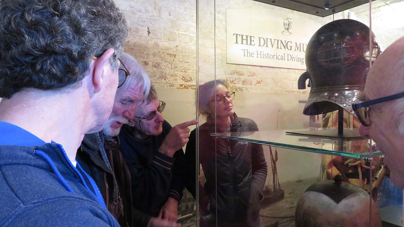 Group of people around the glass cabinet containing the diving helmets looking at the exhibits