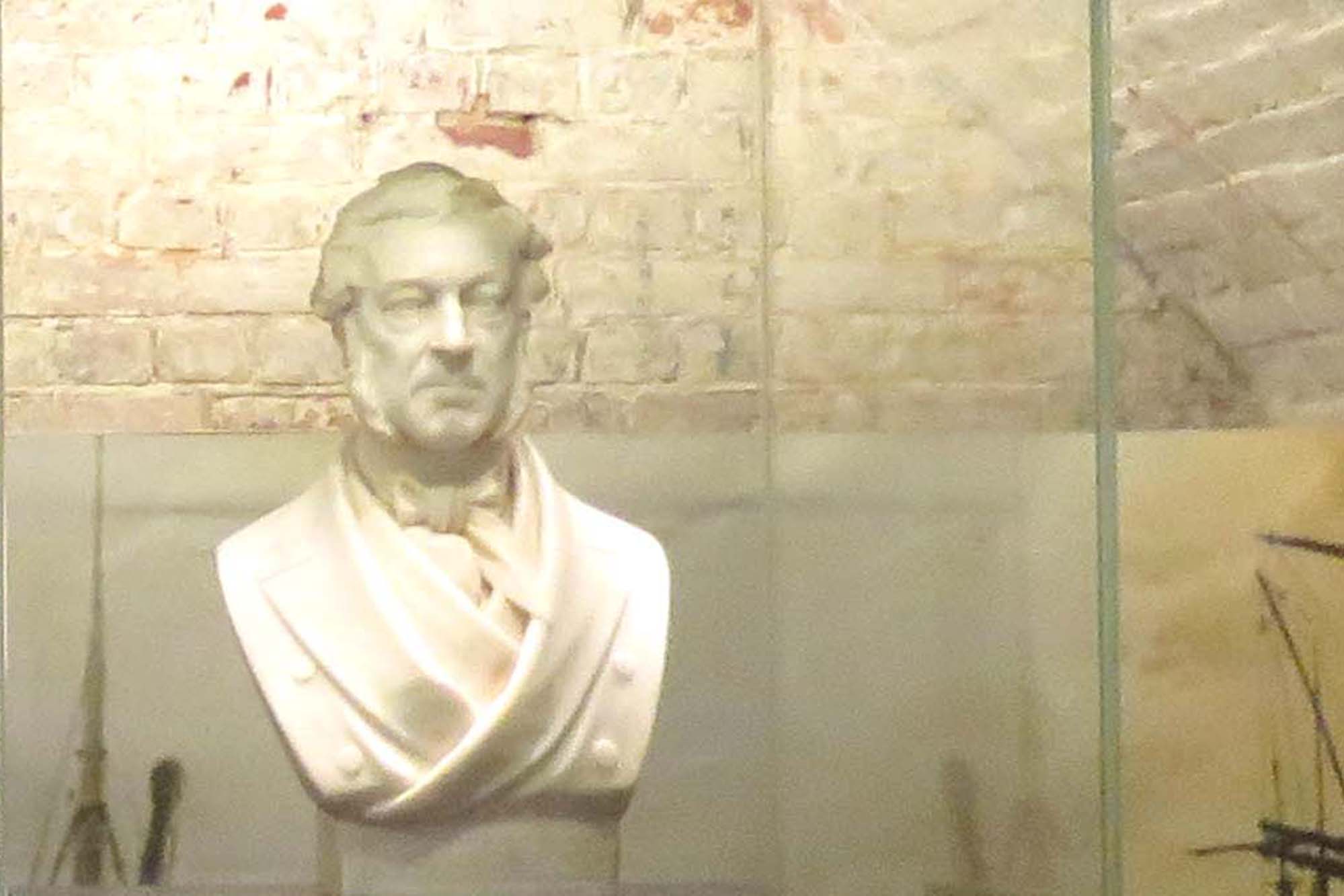 The white marble bust of an elderly Victorian gentleman with sideburns. He is wearing a cravat and overcoat of the period.