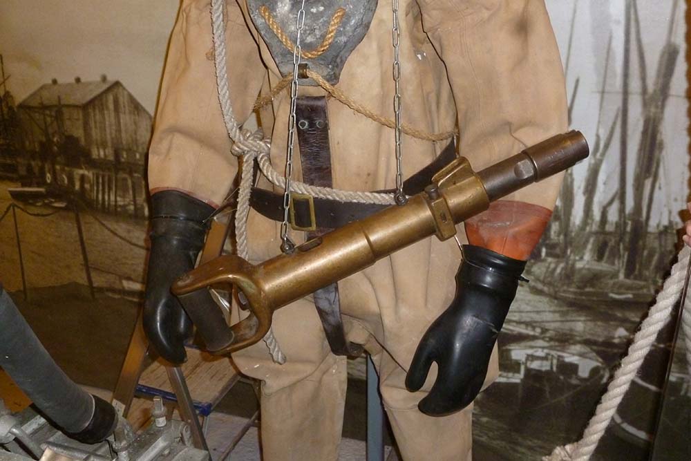 Mid section of a diver in standard dress with a Cox's Bolt Gun slung in front. The diver's gloved right hand holds the handle of the gun which looks like a long brass tube with a handle like a spade's.