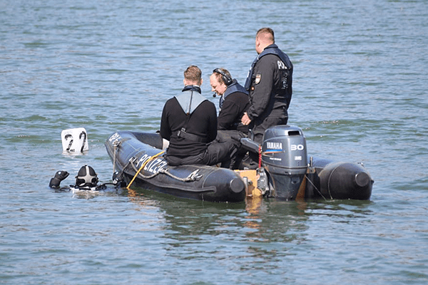 three divers in black dry suits in an inflatable boat with a diver in the water to the left hand side of the boat