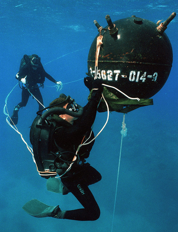 two divers in black suits one in the far background, and one in the foreground reaching up to a spherical black mine