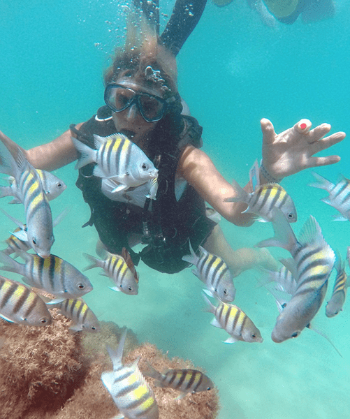 female scuba diver facing the camera surrounded by black and white striped tropical reef fish