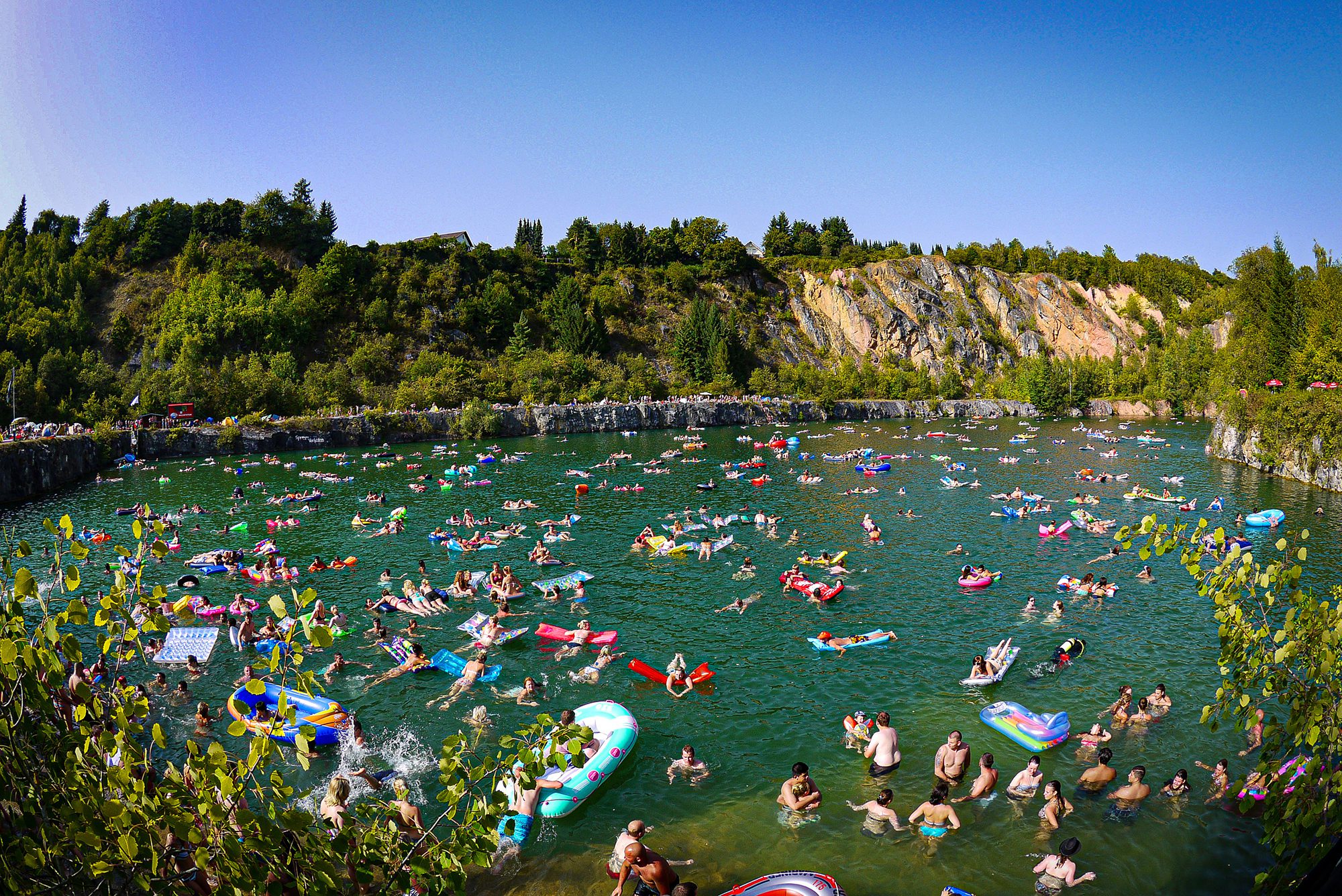 A flooded quarry which is absolutely packed with swimmers and brightly coloured inflatable swim-aids (like rings, lilos and bots) with a scuba diver in the near right foreground