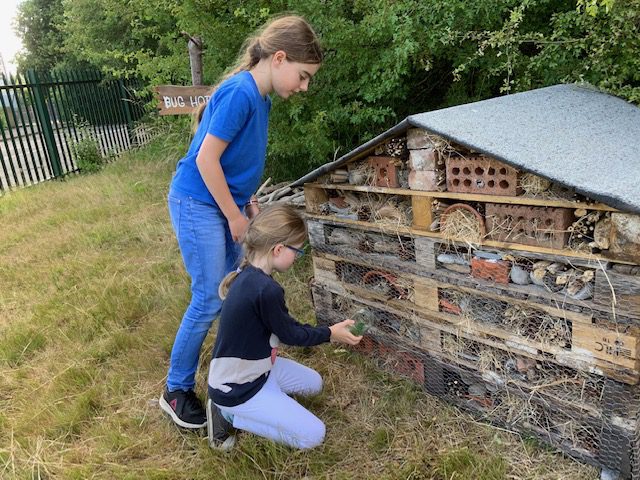 Two young girls, one standing, one kneeling, holding a jam jar containing an insect against a chalet-shaped house made of pallets.