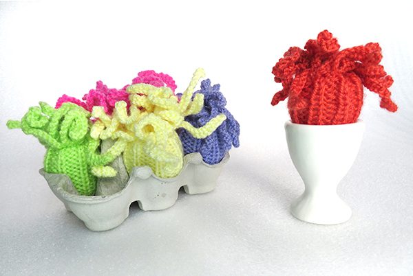 An egg carton containing 5 knitted sea anemones in bright colours with an egg cup on the right with a red knitted sea anemone in itand