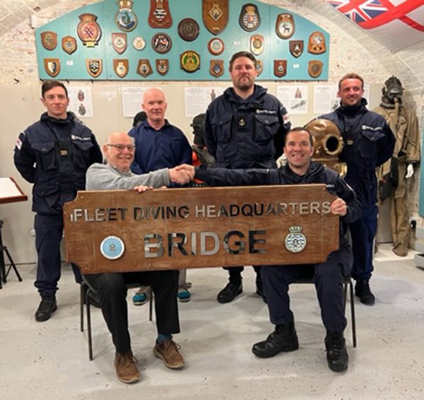 a group of people with two seated in the front row holding each end of a large wooden plaque with the words Fleet Diving HQ Bridge on it.