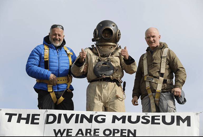 3 men with a diver in the centre dressed in copper helmet standard dress, all standing behind a banner at their feet announcing that the diving museum is open