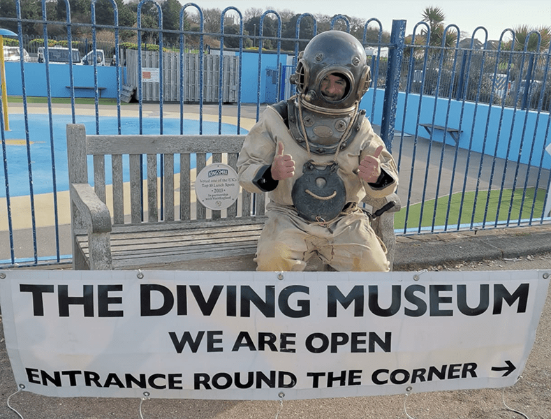 Deep-sea diver in copper helmet seated on a bench with a banner announcing 'The Diving Museum is open'