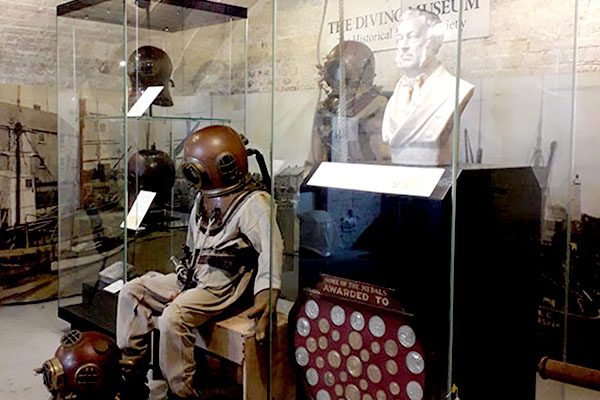 Interior view of the museum - a seated mannequin in standard diving dress between two tall glass cases containing diving helmets and a white marble bust of Augustus Siebe