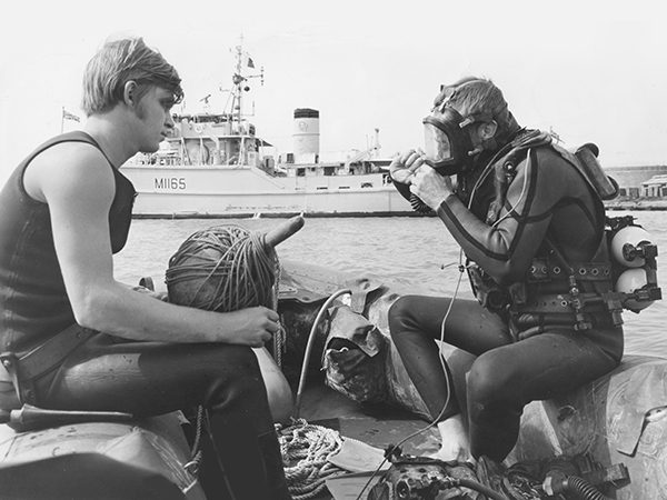 Two diver sitting on the sides of an inflatable boat facing each other while one adjusts his face mask preparing to enter the water
