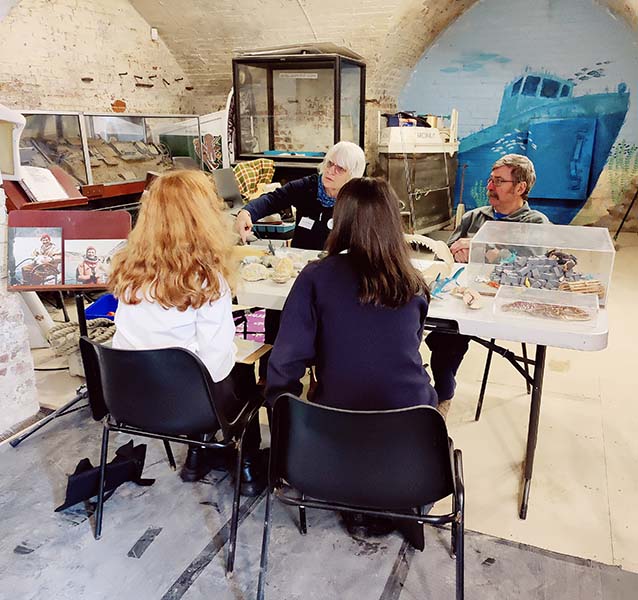 2 adults sitting behind a table covered in artefacts with two children seated (their backs to the camera) in front of the table.