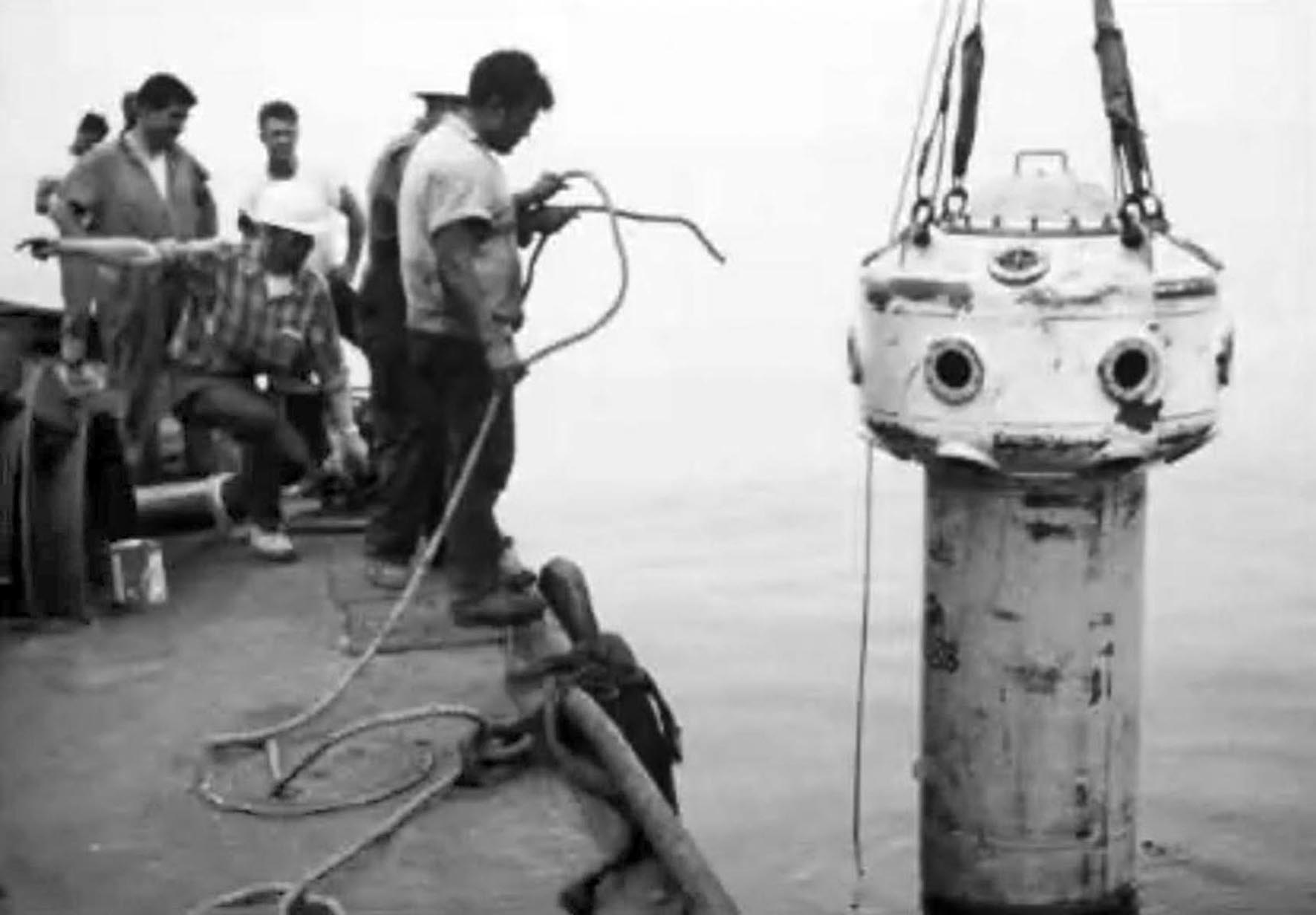 a handful of men standing on the deck of a vessel on the left hand side watching the observation bell being lowered into the sea on the right side of the picture.