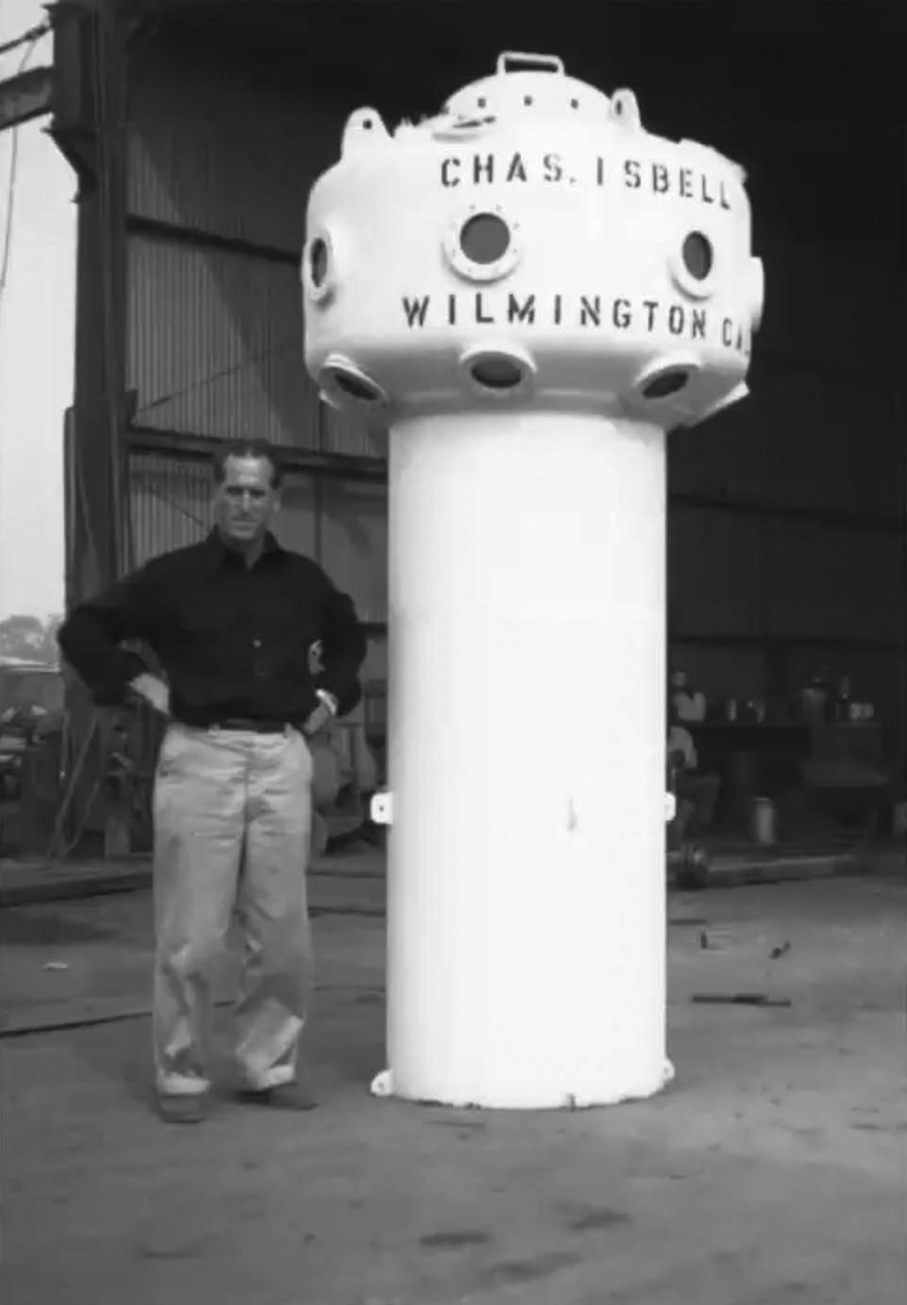 Black and white image of a man facing the camera with hands on hips. On his left is an observation bell/chamber painted white with the words Chas. Isbell, Wilmington CA. painted around the top part of the bell.