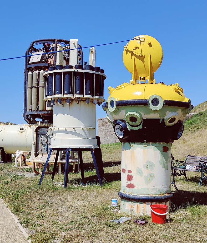A view of 3 diving bells outside the museum with the one in the foreground, an observation bell, with it's lid open and top part being painted in yellow.